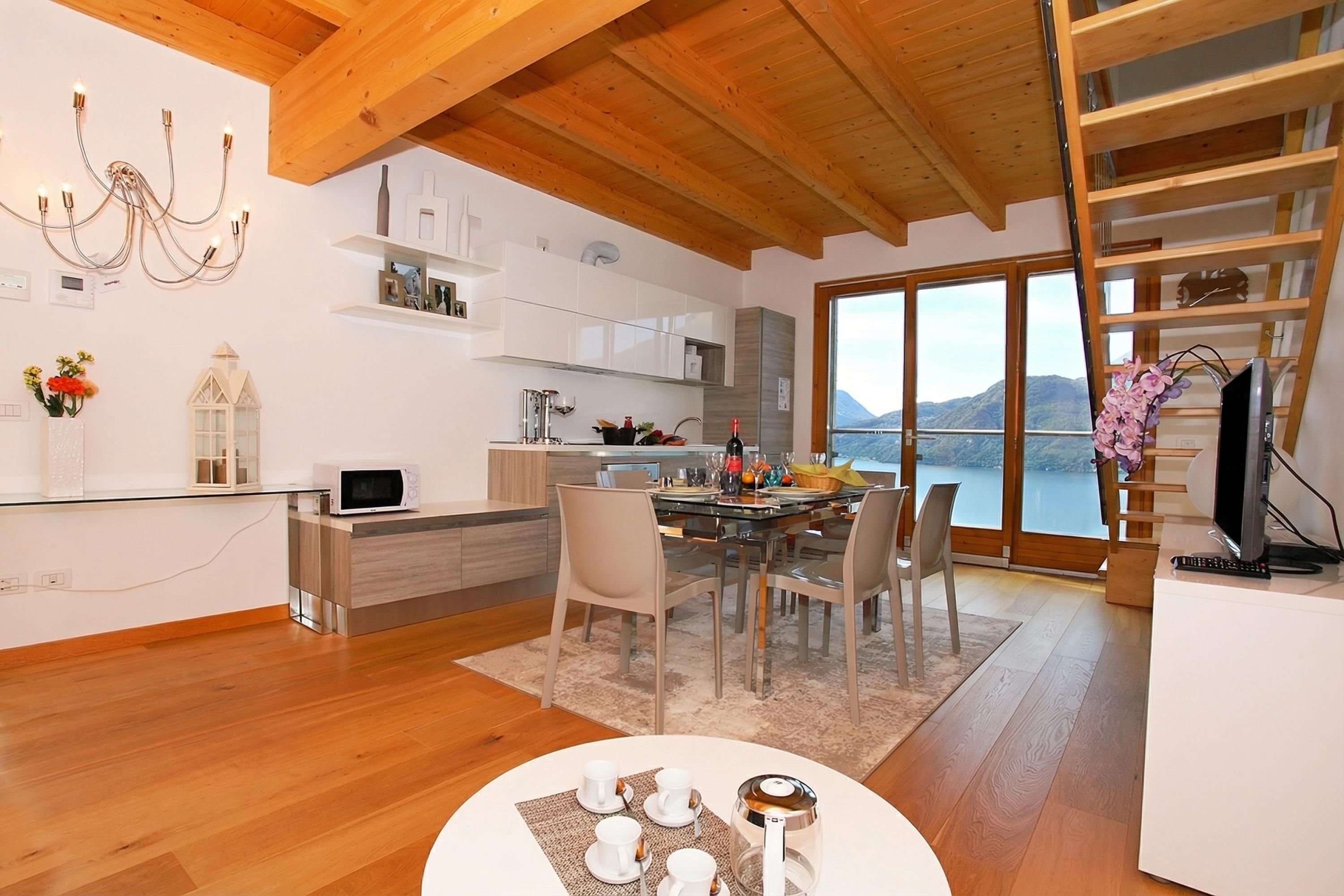 Una cocina moderna con armarios blancos, un comedor con mesa de vidrio y vistas a un lago a través de grandes ventanales.