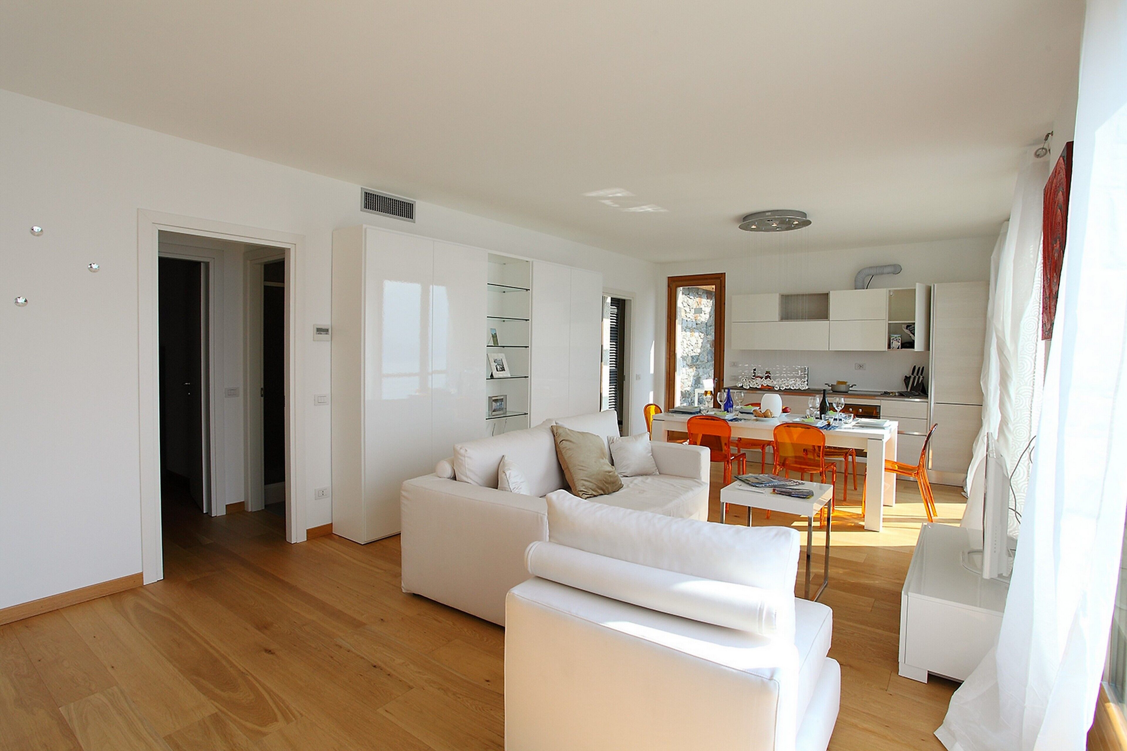 A modern living room with a white sofa, a dining area with orange chairs, and a kitchen in the background.