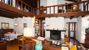 A spacious living room with a high ceiling, wooden beams, a fireplace, a dining area with a table set for two, and a collection of bookshelves.
