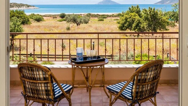 Un balcón con sillas de mimbre, una mesita y una bandeja con vasos y una jarrón, con vistas a un paisaje costero con montañas al fondo.