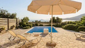Zona de piscina con sillas de descanso y una sombrilla, rodeada de baldosas de piedra y follaje.
