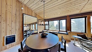 A modern dining area with a wooden table and chairs, large windows, and a stone wall. - Spacious 3 Bedroom Condo with Loft - Hike, Bike, Swim, Ski! by RedAwning (Mammoth Lakes)