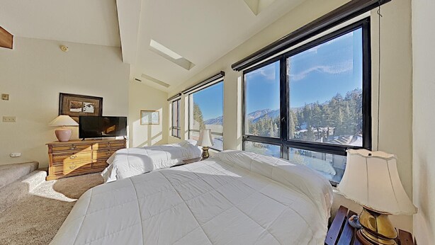 A bedroom with a large bed, a dresser, a TV, and a view of mountains through large windows. - Slopeside 1849 Condos - Comfortable 3 BR Condos with Full Kitchens (Mammoth Lakes)
