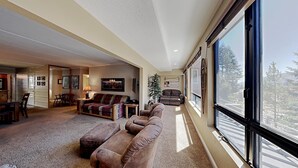 A spacious living room with a high ceiling, large windows offering a view of trees, a fireplace, and a cozy seating area. - 2 Bedroom Condo Just Steps Away From Summer Recreational Activities by RedAwning (Mammoth Lakes)