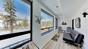 Una habitación con un ventanal grande que ofrece vistas a un paisaje nevado y árboles, un sofá de cuero negro, una mesita y un adorno de pared.