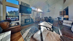 A spacious living room with a stone fireplace, leather sofas, a dining area, and a large window offering a view of trees. - Large Upgraded 4 Bedroom Condo in Mammoth! Easy access To Hot Tubs, Pool, Sauna by RedAwning (Mammoth Lakes)