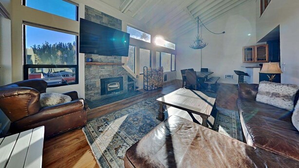 A spacious living room with a stone fireplace, leather sofas, a dining area, and a large window offering a view of trees. - Large Upgraded 4 Bedroom Condo in Mammoth! Easy access To Hot Tubs, Pool, Sauna by RedAwning (Mammoth Lakes)