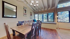 A dining area with a wooden table and chairs, a chandelier, and a large window with a view of trees. - Family Friendly Spacious Mountain 4 Bedroom Condo - Closest to Canyon Lodge! by RedAwning (Mammoth Lakes)