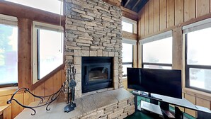 A stone fireplace with a TV on a stand, wooden walls, and large windows.