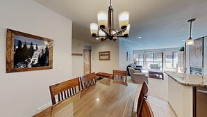 A dining area with a wooden table and chairs, a chandelier, and a large window with a mountain view. - Stay mountainside! One Bedroom Condo Just Steps to the Mountain and 1 Mile to the Village at Mammoth by RedAwning (Mammoth Lakes)