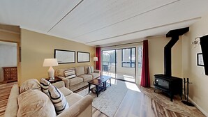 A spacious living room with a sofa, a coffee table, and a fireplace. There are framed pictures on the wall and a window with curtains.