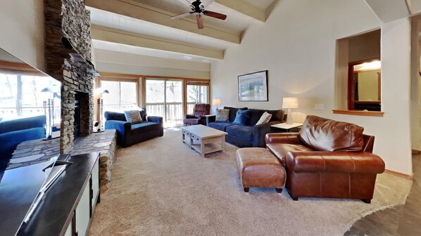 A spacious living room with a stone fireplace, a ceiling fan, and large windows allowing natural light. - Beautiful Mountainside 4 BR Plus Loft/4 Bath Home in Mammoth! by RedAwning (Mammoth Lakes)