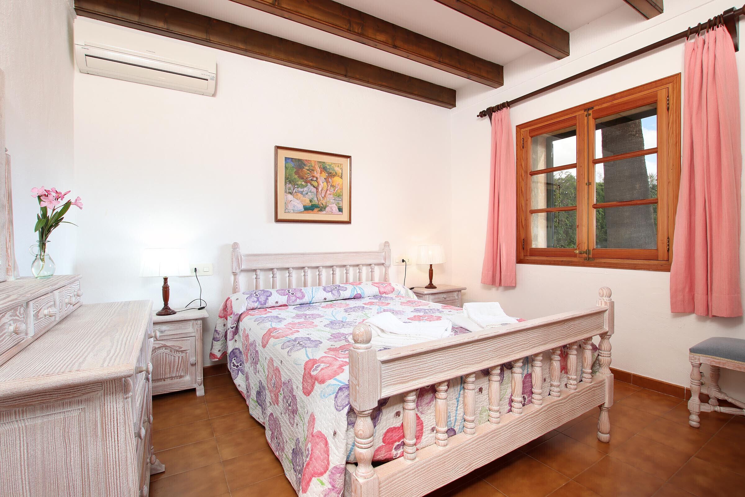 Een slaapkamer met een houten bed, een nachtkastje, een dressoir, een raam met gordijnen en een schilderij aan de muur.