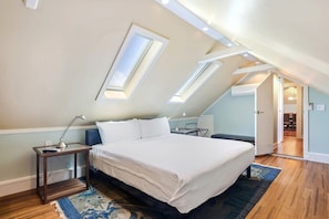 Een slaapkamer met een bed, nachtkastjes, een bureau en een stoel. Er zijn dakramen en een deur die naar een andere kamer leidt.