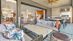 A spacious living area with a sofa, coffee table, and dining table. There is a kitchen with a grill and a ceiling fan.
