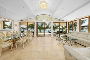 A spacious living room with a glass table, beige sofas, and a view of palm trees and the sea.