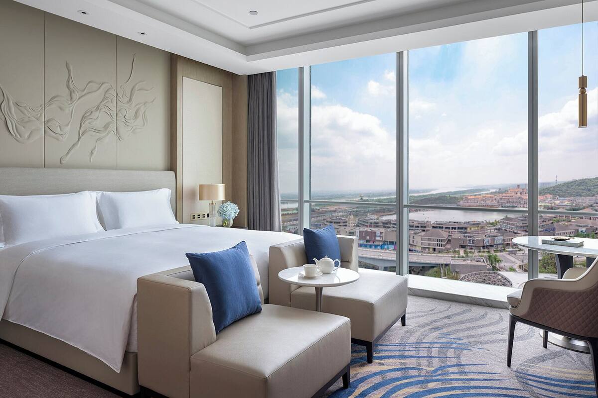 A hotel room with a large bed, two armchairs, a small table, and a view of a cityscape through large windows.
