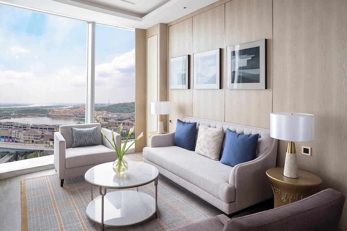 A modern hotel room with a large window offering a city view, a sofa set, a coffee table, and two side tables with lamps.