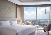A modern hotel room with a large bed, a view of a cityscape, a sitting area with a table and chair, and a decorative wall piece.
