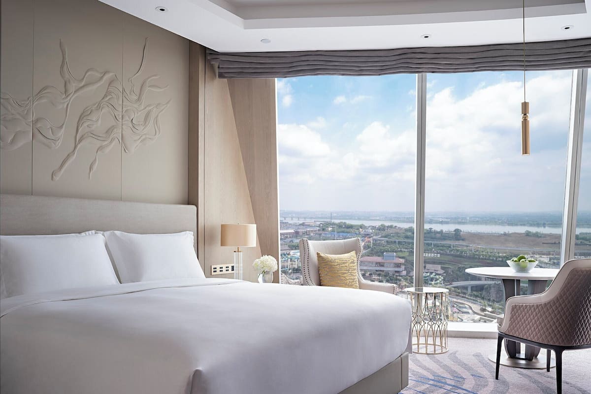 A modern hotel room with a large bed, a view of a cityscape, a sitting area with a table and chair, and a decorative wall piece.