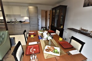 Een eetruimte met een lange tafel gedekt voor vier personen, voorzien van rode placemats, een vaas met bloemen en een bord eten.
