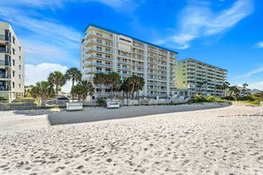 A beachfront hotel with multiple floors, balconies, and a sandy beach in the foreground.