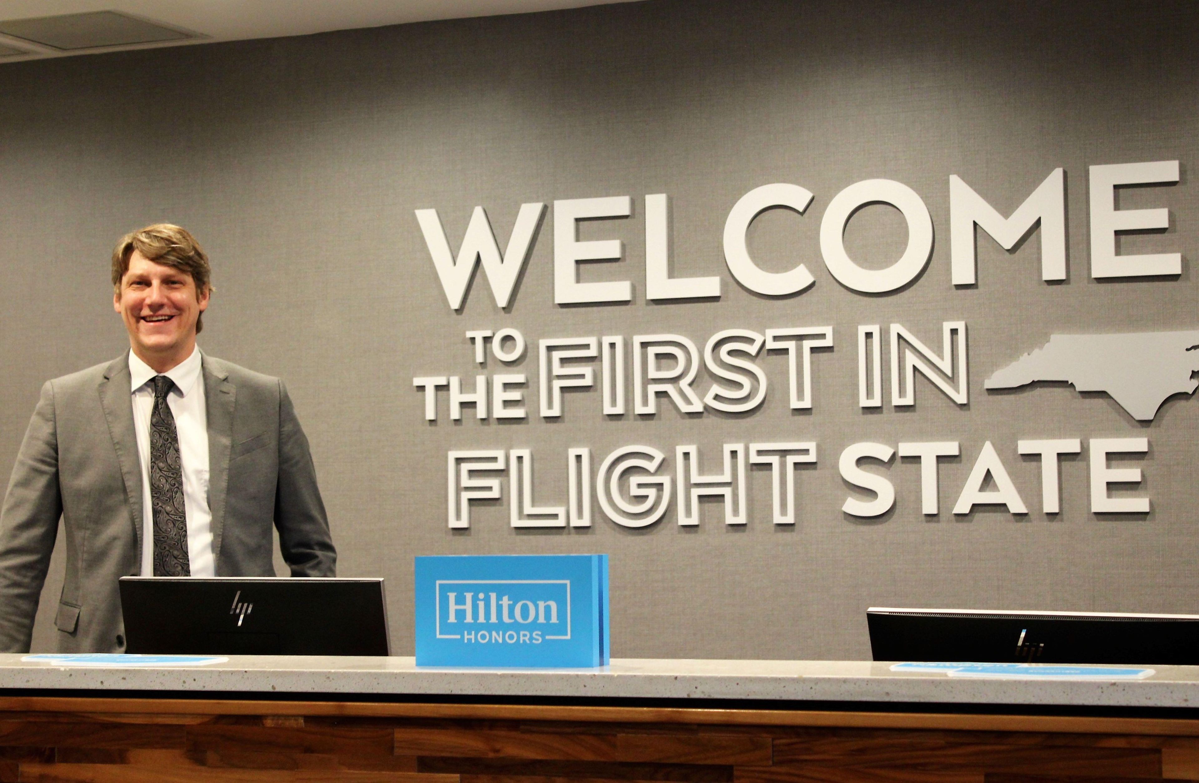 A man standing at a reception desk with a 'Welcome to the First in Flight State' sign in the background.
