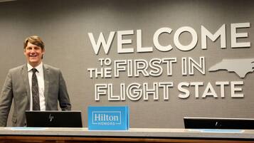A man standing at a reception desk with a 'Welcome to the First in Flight State' sign in the background.