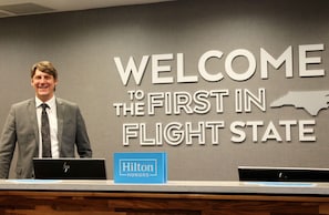 A man standing at a reception desk with a 'Welcome to the First in Flight State' sign in the background. - Hampton Inn Lexington, NC (Lexington)