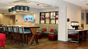 Een hotellobby met een lange tafel, meerdere stoelen, een bank, een koffiehoek en een televisiescherm.