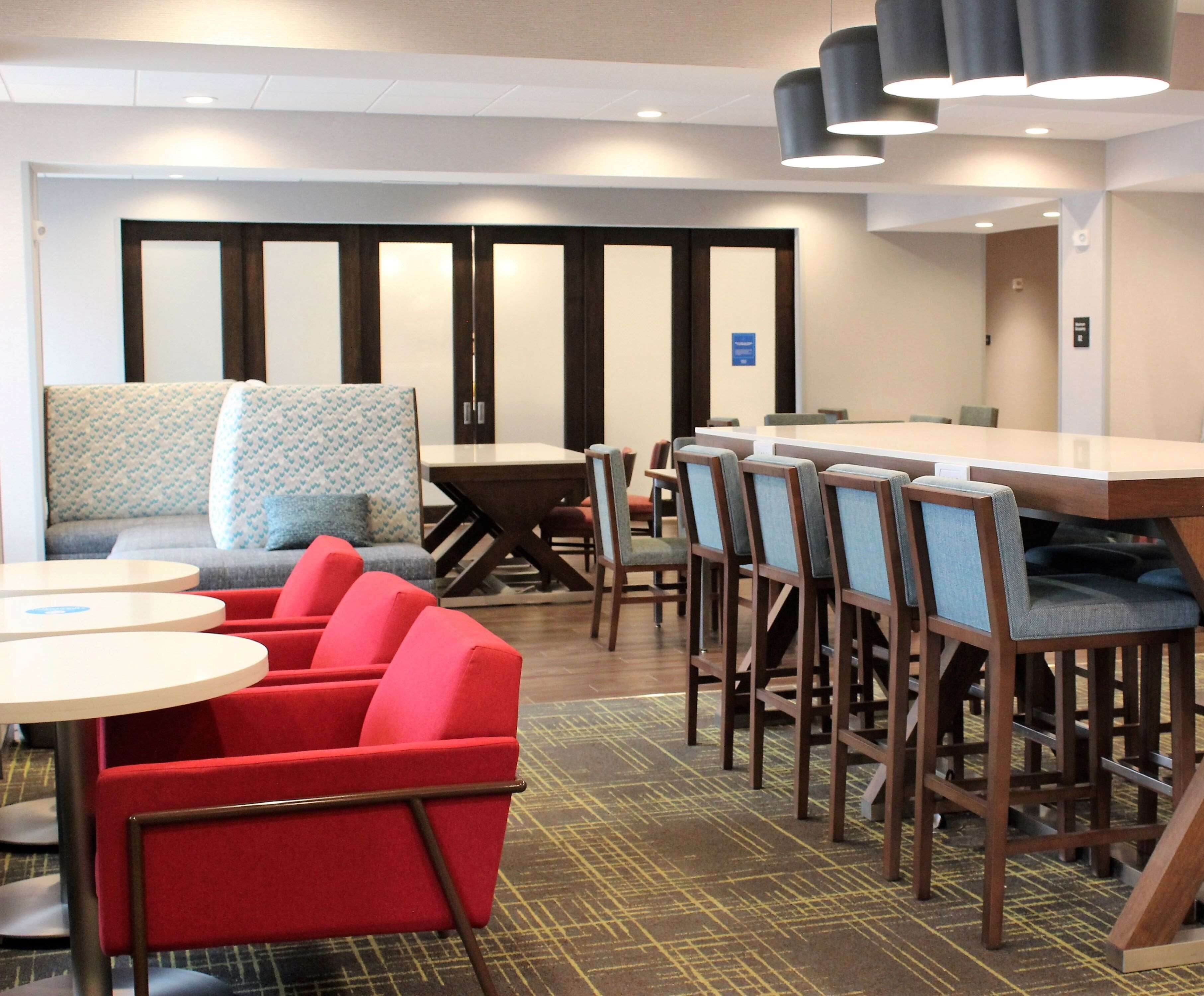 Un hall de hotel con un área de descanso que incluye sillas rojas, un sofá y una mesa. Hay varias mesas y sillas dispuestas para comer o celebrar reuniones.