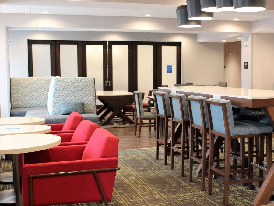 Un hall de hotel con un área de descanso que incluye sillas rojas, un sofá y una mesa. Hay varias mesas y sillas dispuestas para comer o celebrar reuniones.