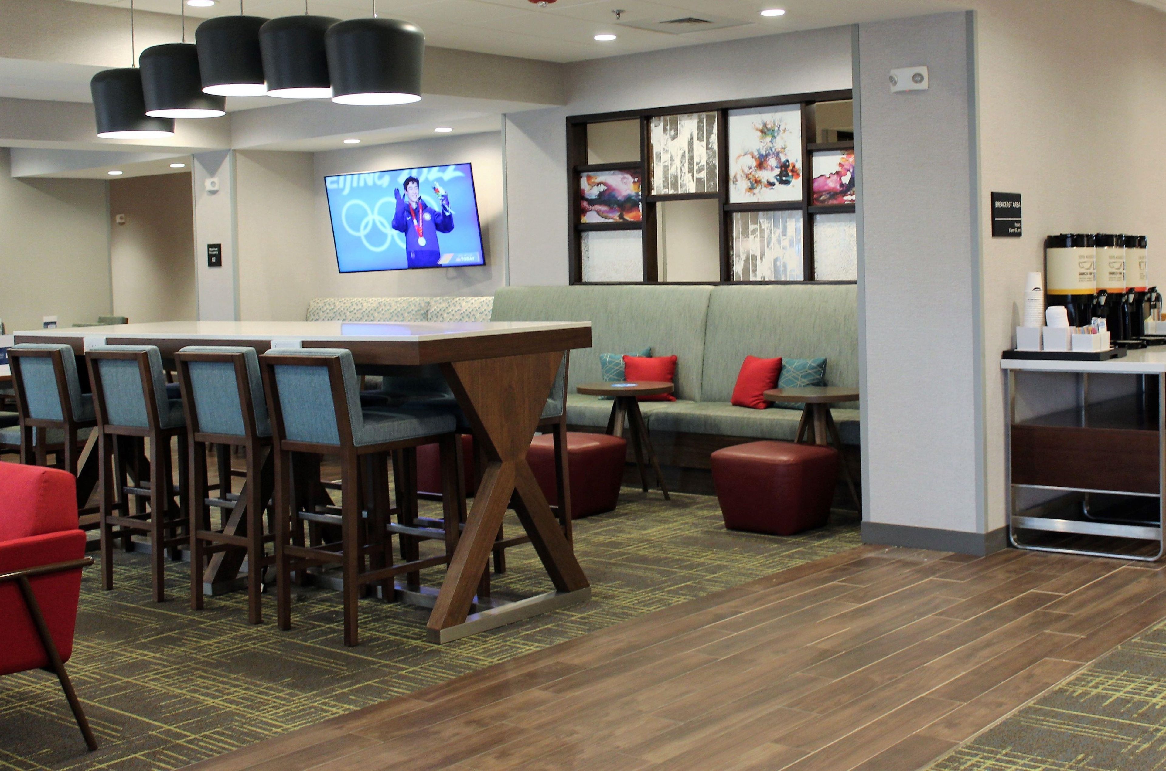 A hotel lobby with a long table, several chairs, a sofa, and a television displaying the Olympic rings.