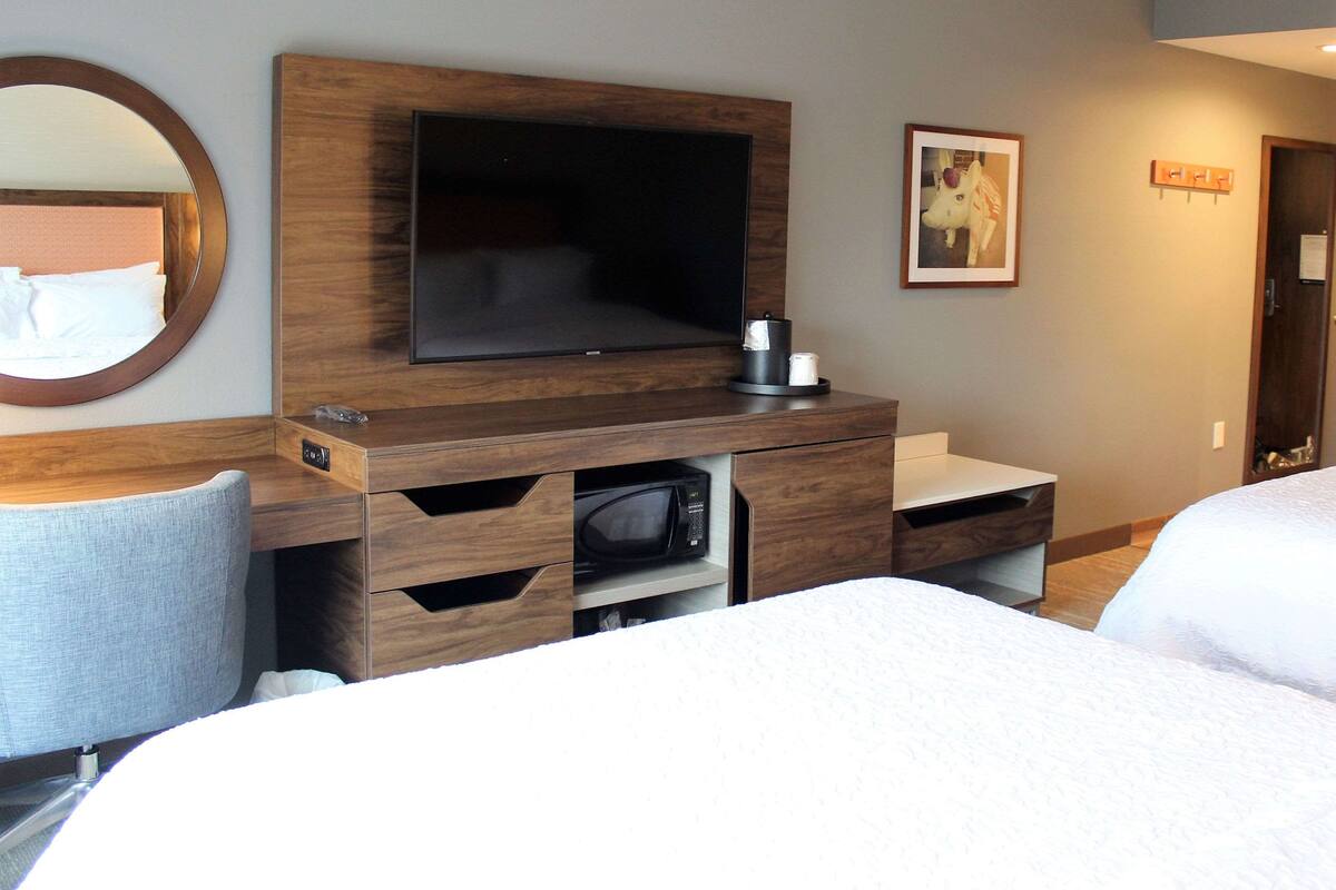 A hotel room with a large flat-screen TV mounted on a wooden cabinet, a microwave, and a small coffee maker. There are two beds with white linens and a round mirror on the wall.