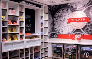 Una sección de aperitivos y bebidas con diversas opciones empaquetadas, un cartel de pizarra y un expositor refrigerado.