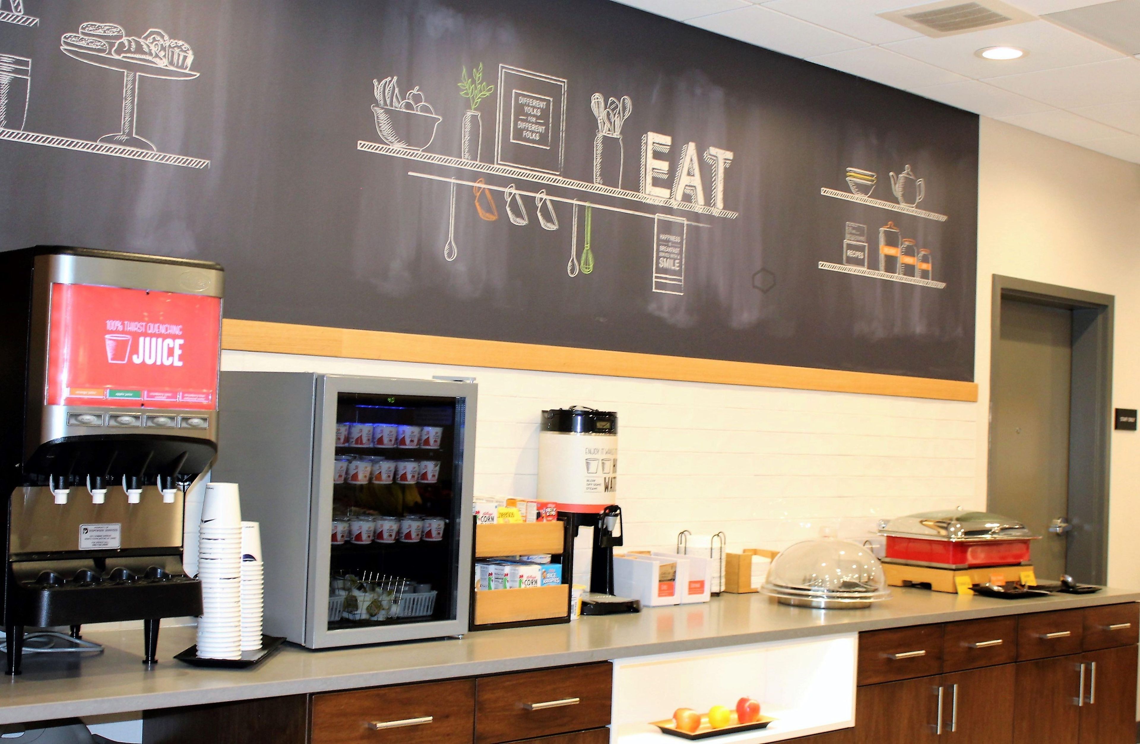 A self-service beverage station with a coffee machine, a refrigerator stocked with drinks, and a chalkboard wall with food-related drawings.