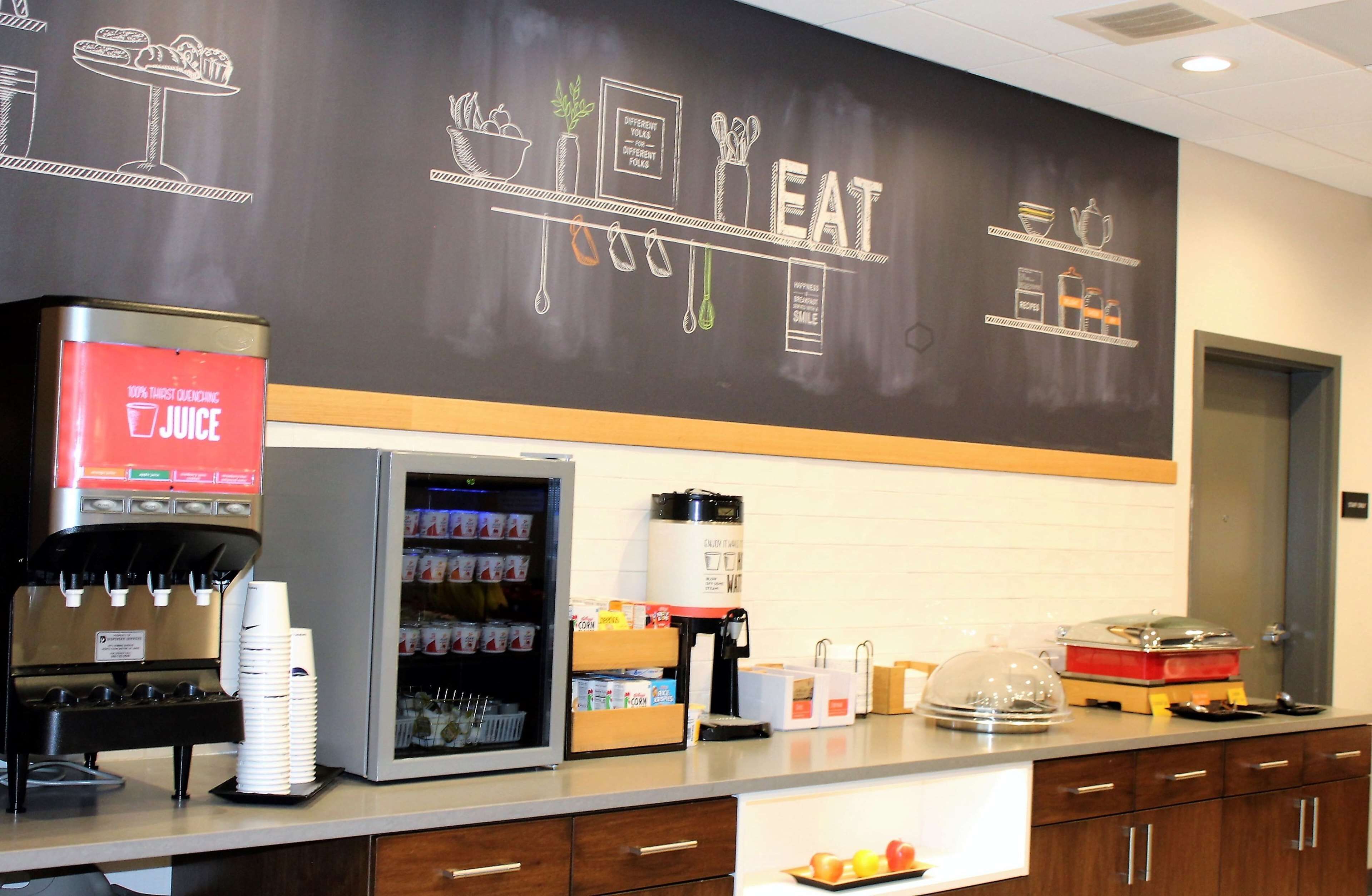 Una estación de café con un dispensador de jugo, un refrigerador lleno de bebidas y un menú en una pizarra.