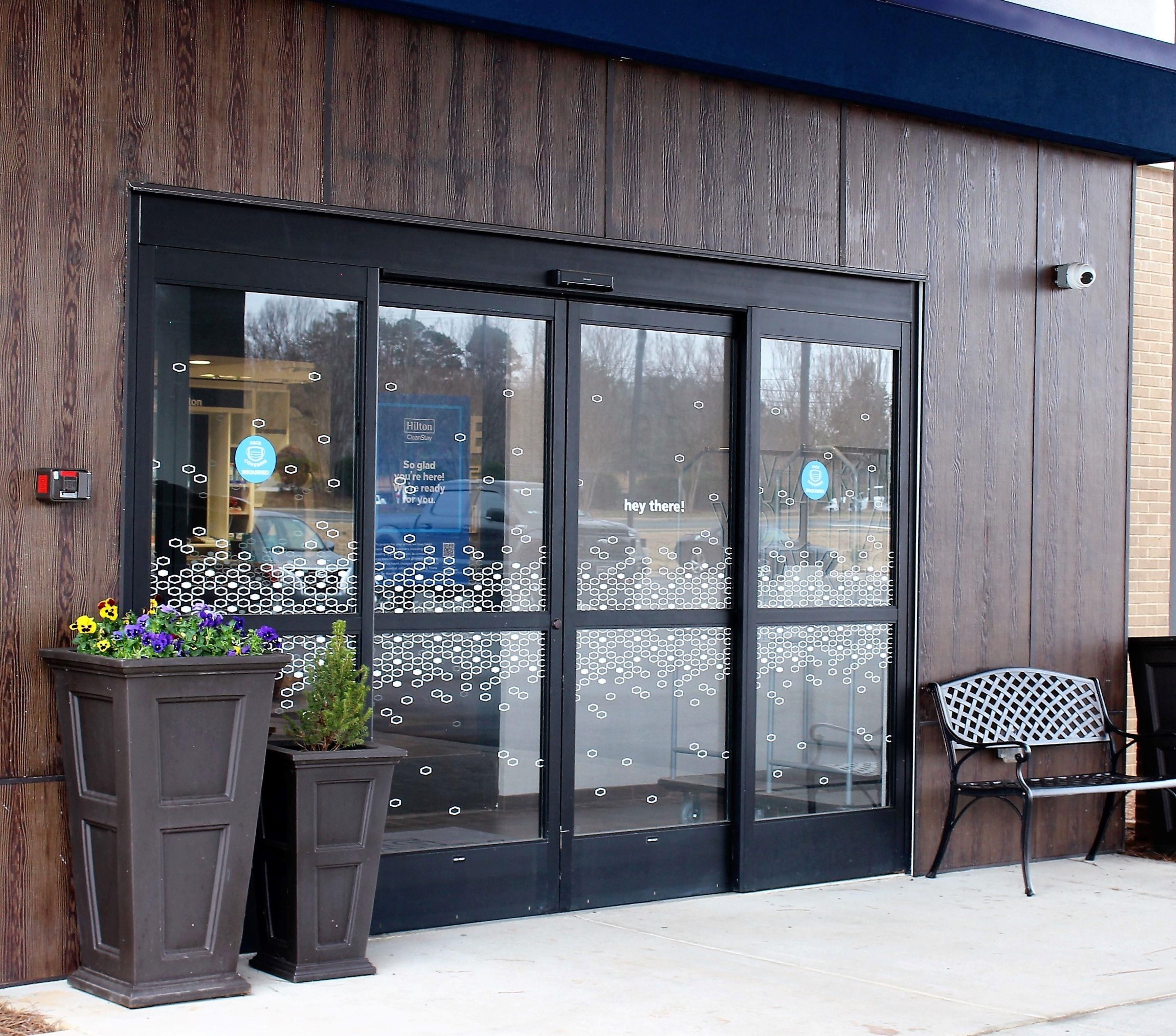 A hotel entrance with a large glass door, a potted plant with flowers, and a metal chair outside.