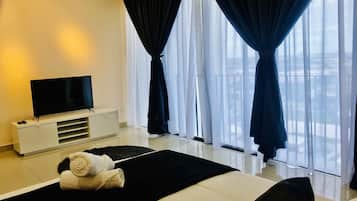 A hotel room with a bed, a TV on a white cabinet, and a view of the city through sheer curtains.