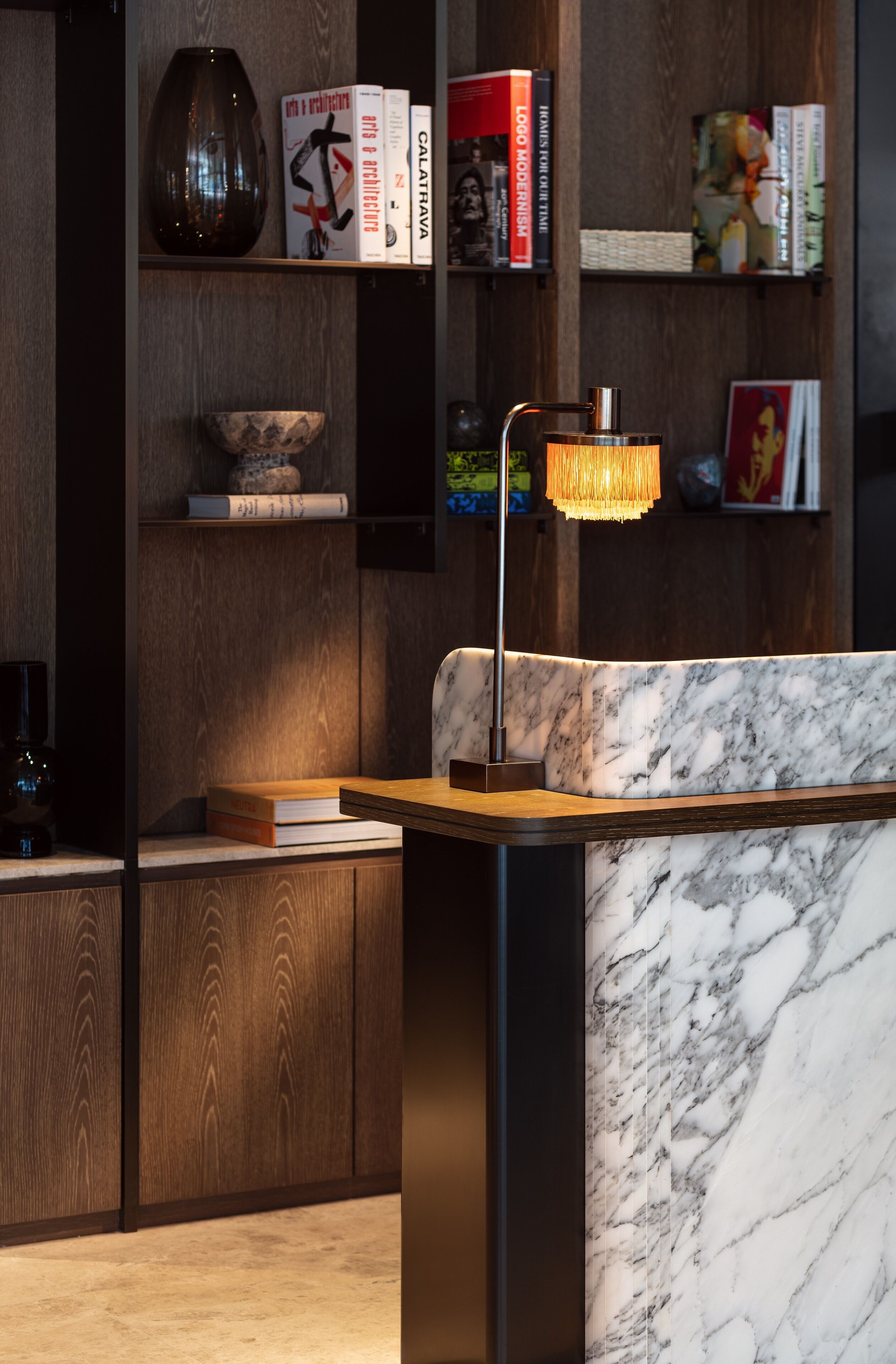A modern interior with a marble reception desk, wooden shelves with books, and a decorative lamp.