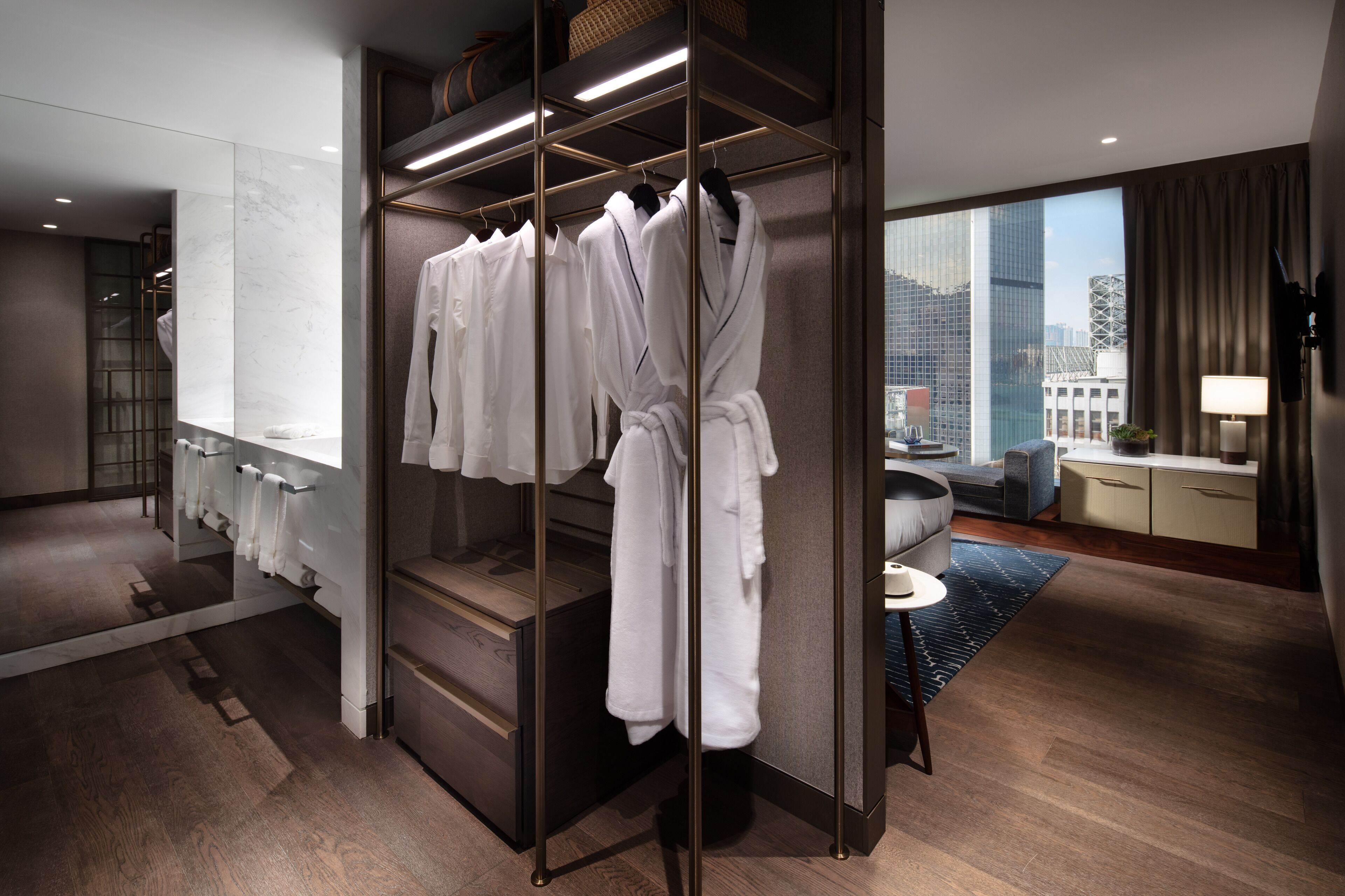 Habitación de hotel moderna con vestidor que contiene batas blancas, un espejo grande y vistas a la ciudad.
