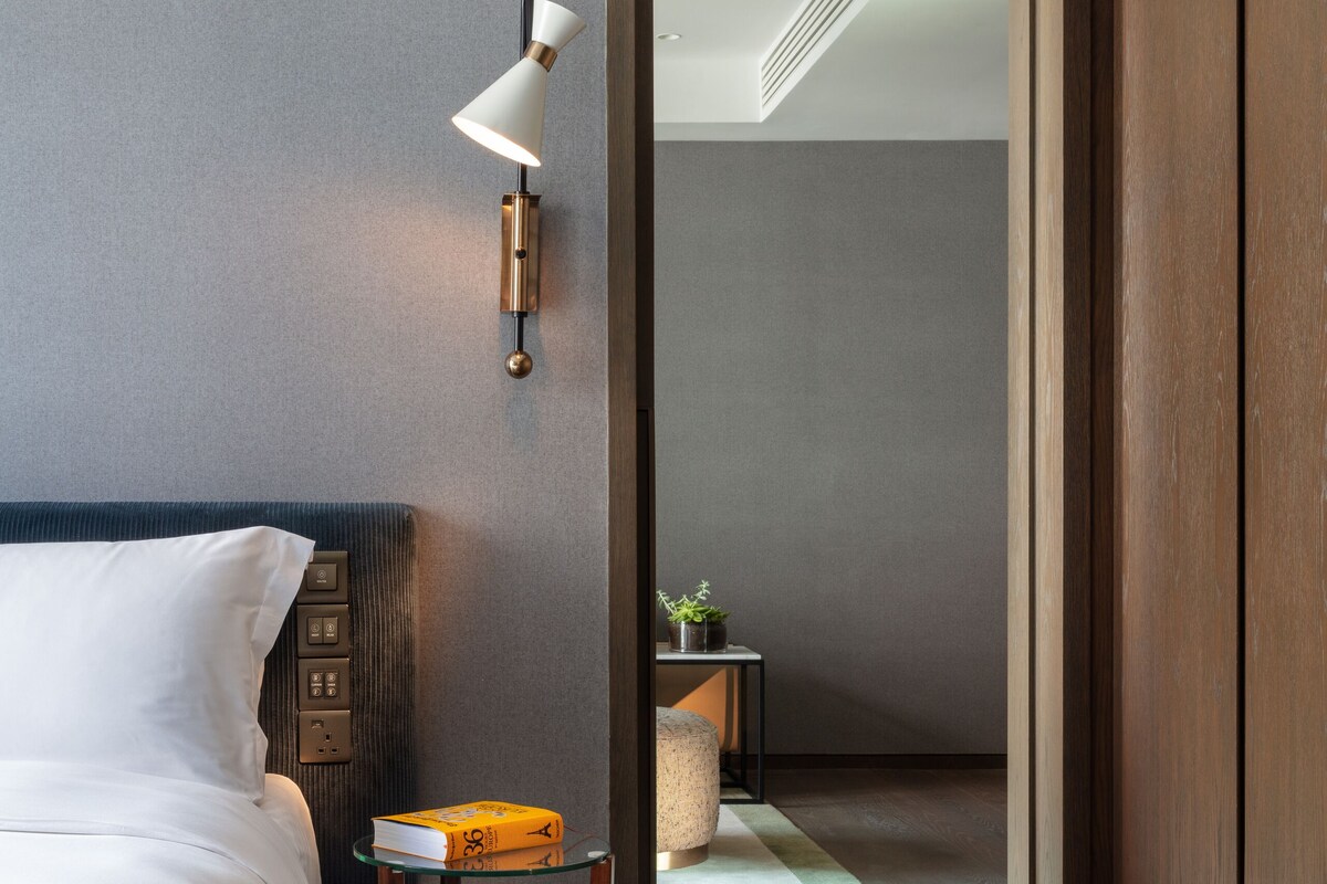 A modern hotel room with a neatly made bed, a bedside table with a lamp, and a small side table with a plant.