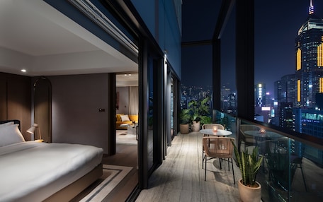 A modern hotel room with a balcony, a bed, a seating area, and a city view.