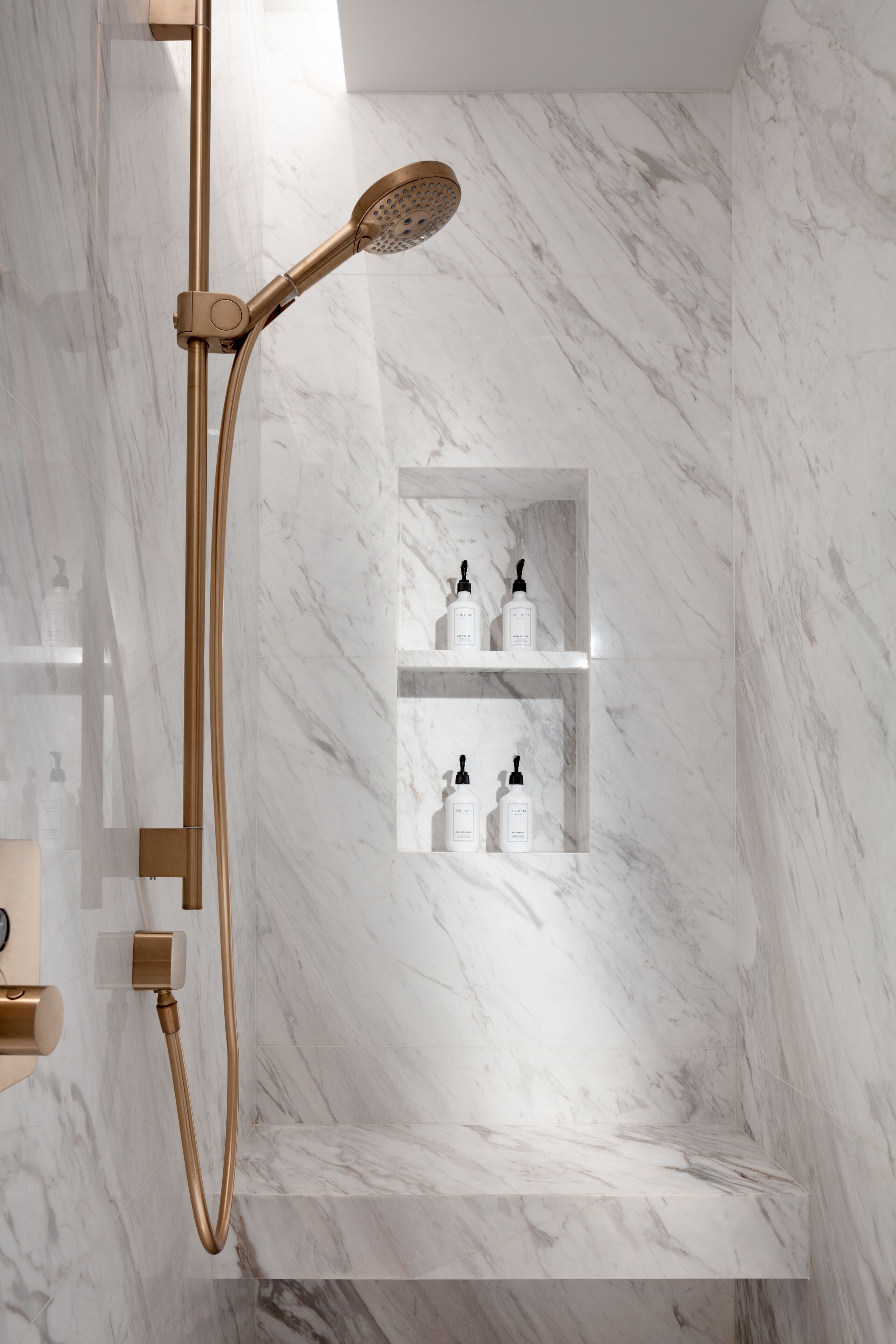 A modern bathroom with a marble floor, a gold showerhead, and built-in shelves with toiletries.