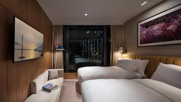 Habitación de hotel moderna con dos camas, un sofá, una lámpara de lectura y un amplio ventanal con vistas a un paisaje urbano.