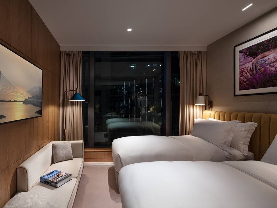Habitación de hotel moderna con dos camas, un sofá, una lámpara de lectura y un amplio ventanal con vistas a un paisaje urbano.