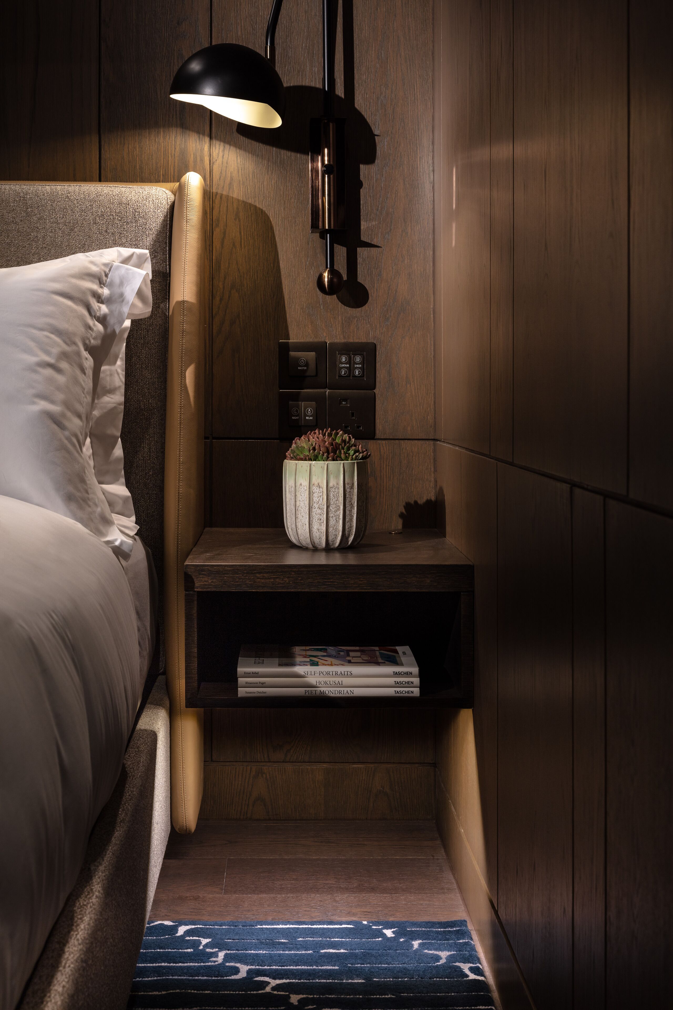 A neatly made bed with white linens, a bedside lamp, a small wooden nightstand with a potted plant, and a magazine rack.