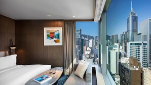A modern hotel room with a large window offering a city view, a neatly made bed, a small seating area, and a framed artwork on the wall. - The Hari Hong Kong (Hong Kong)