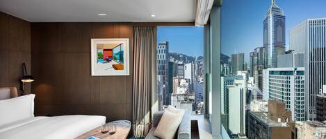 A modern hotel room with a large window offering a city view, a neatly made bed, a small seating area, and a framed artwork on the wall.