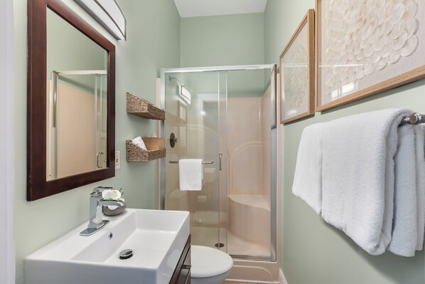 A bathroom with a glass-enclosed shower, a white sink, and a toilet. There are wicker baskets on the wall and a white towel hanging on the door.